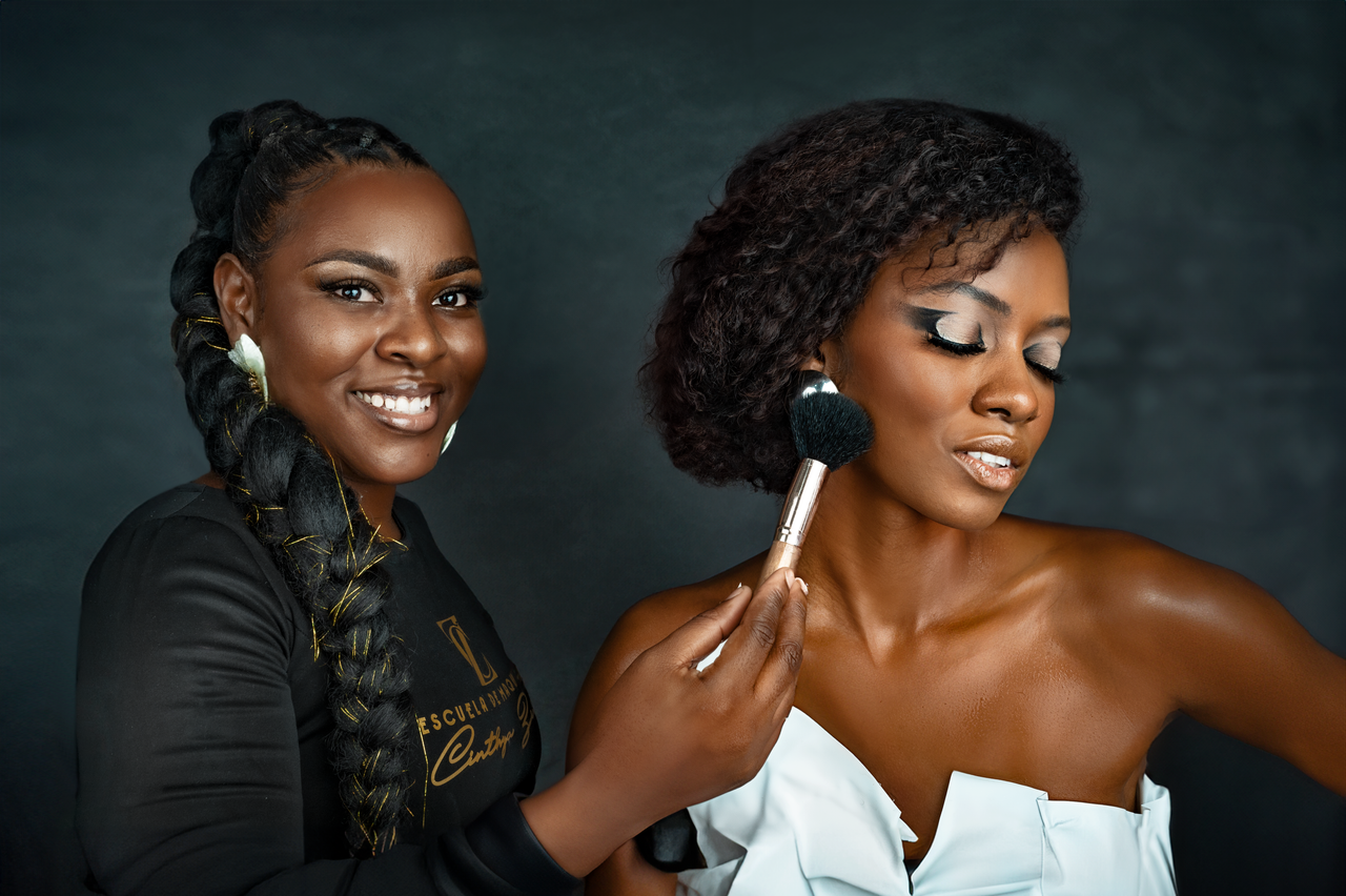 A fashion portrait of two women in a studio with one applying makeup to the other smiling.