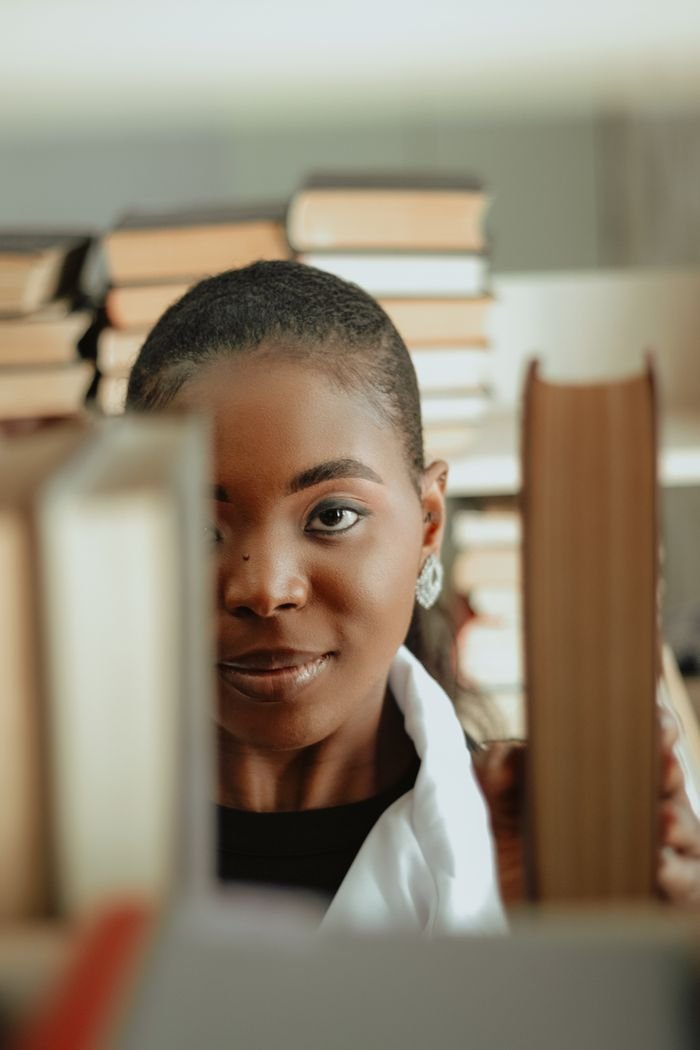 Elegant portrait of a woman peeking through bookshelves in a library setting.